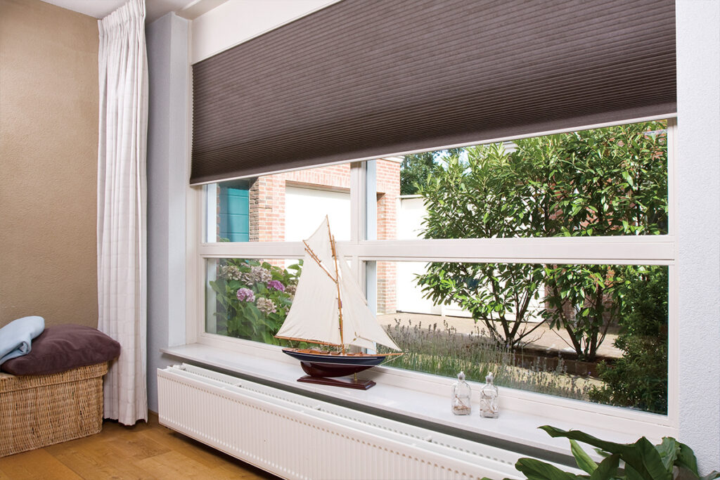 Stylish blinds covering the top half of a window. The windowsill contains a model sailing ship and two candles. Trees are outside the window.